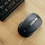 Souris sans fil noire posée sur un bureau en bois, à côté d'un clavier.