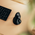 Souris verticale noire posée sur un tapis de souris beige, à côté d'un clavier noir.