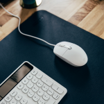 Souris filaire blanche connectée à un clavier blanc, posés sur un bureau en bois clair avec un tapis noir.