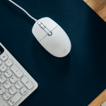 Souris filaire blanche posée sur un tapis de bureau noir, à côté d'un clavier assorti.