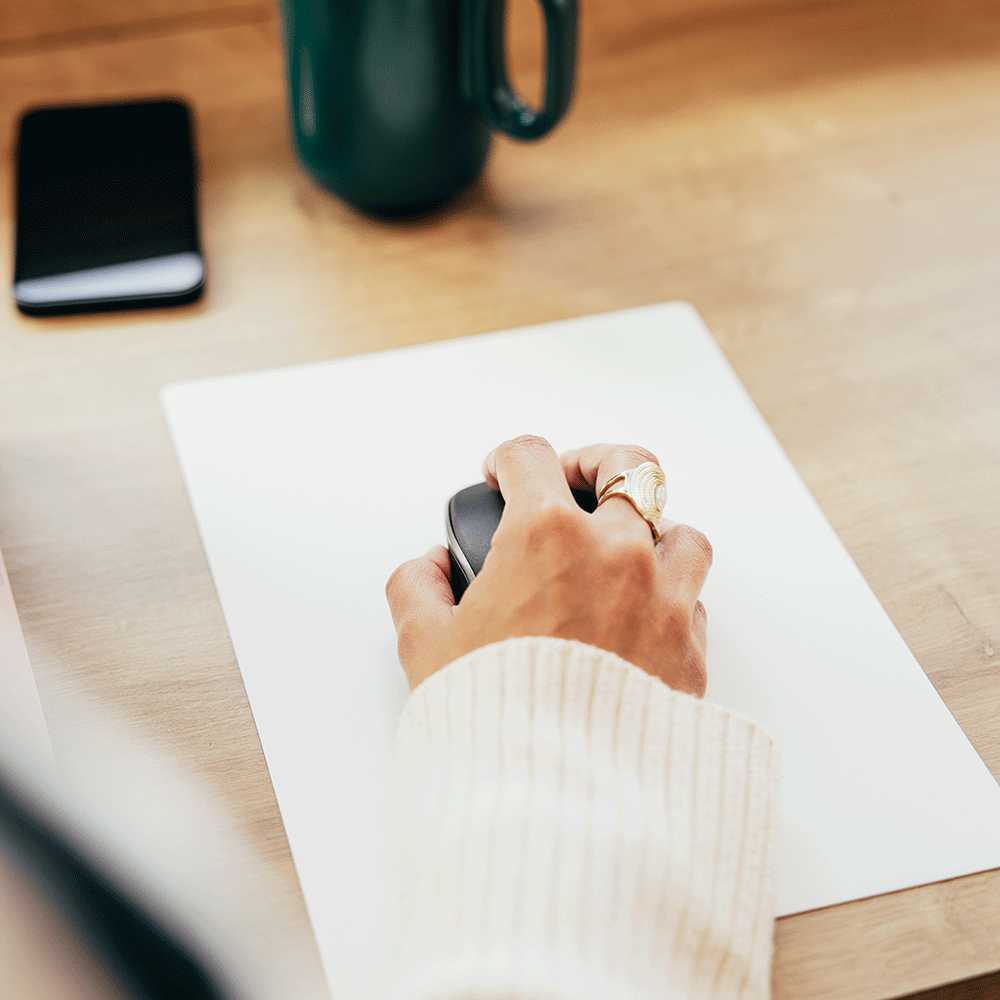 Main utilisant une souris sur un tapis blanc, avec une tasse verte posée sur un bureau clair.