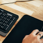 Main utilisant une souris sur un tapis de souris noir, posé sur un bureau avec un clavier à proximité.